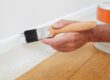 Close up of a man painting the baseboard of a home in white paint.