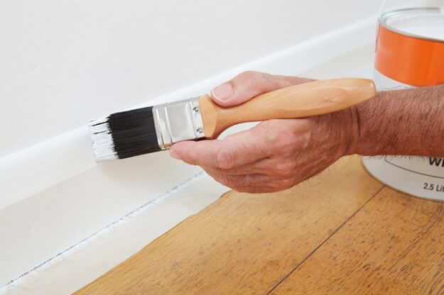 Painting Special Surfaces Close up of a man painting the baseboard of a home in white paint.